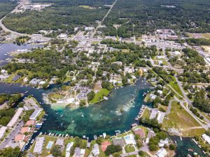 Aerial view of a small coastal town with residential homes, lush greenery, and a clear water bay filled with boats.