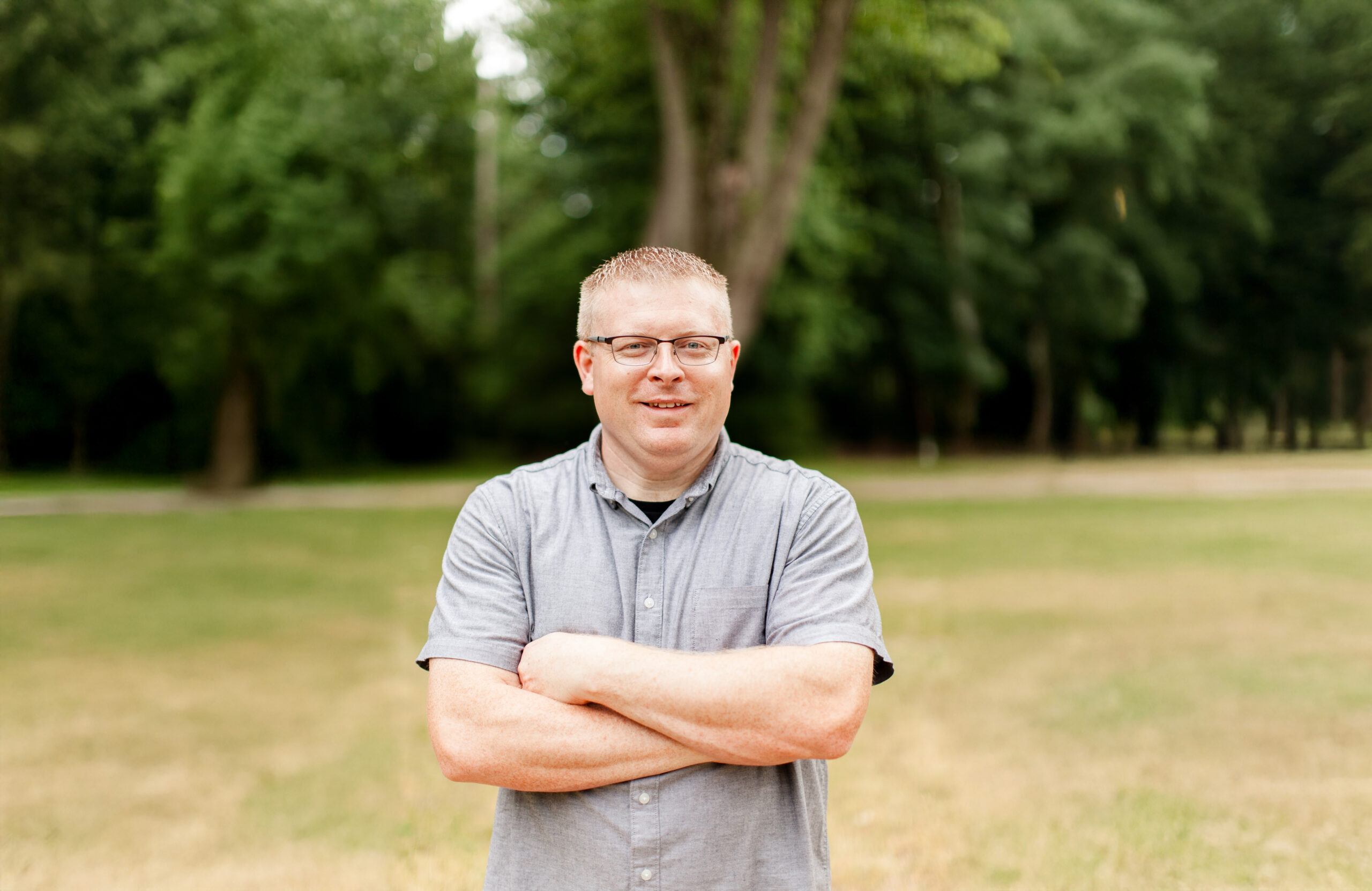 John Leutermann, wearing glasses and a gray shirt, stands outdoors with arms crossed.